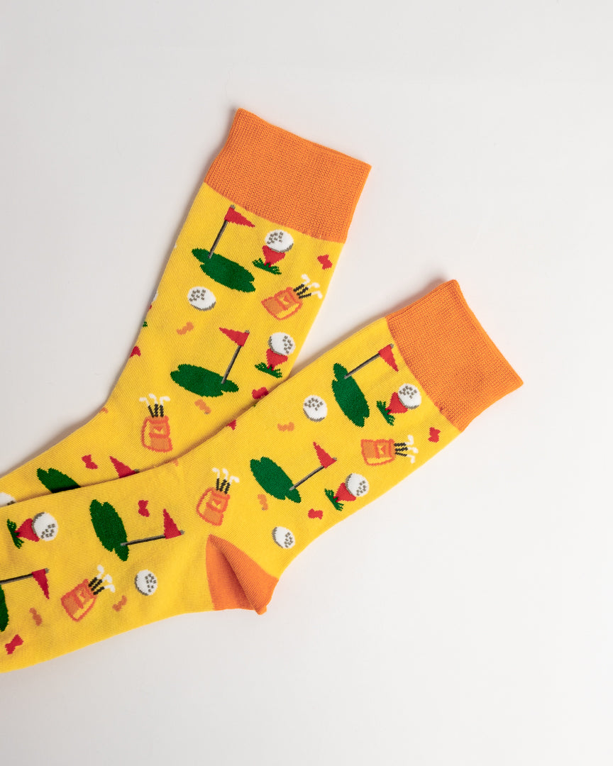 A pair of bright yellow crew socks with orange cuffs and toes, featuring a playful golf course pattern with green tees, flags, and golf balls, displayed on a clean white background.