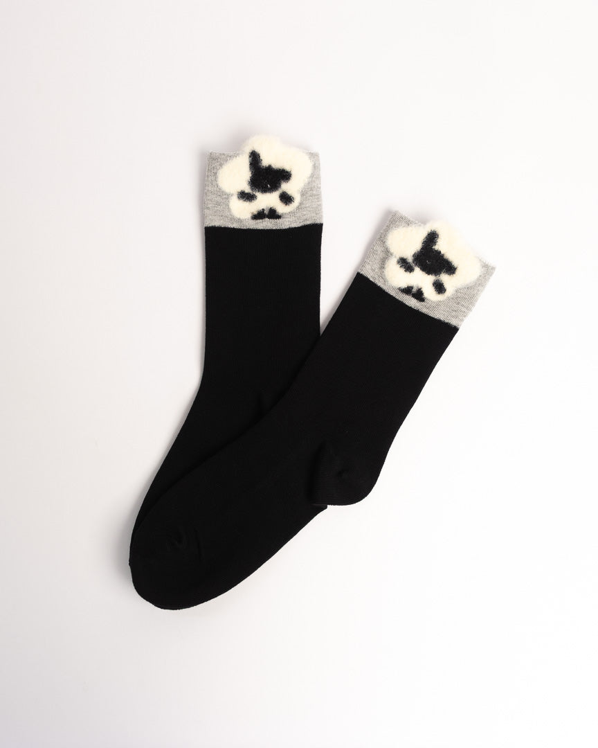 A pair of black crew socks with a gray ribbed cuff and a white fuzzy pom-pom detail shaped like a black paw print, displayed on a clean white background.
