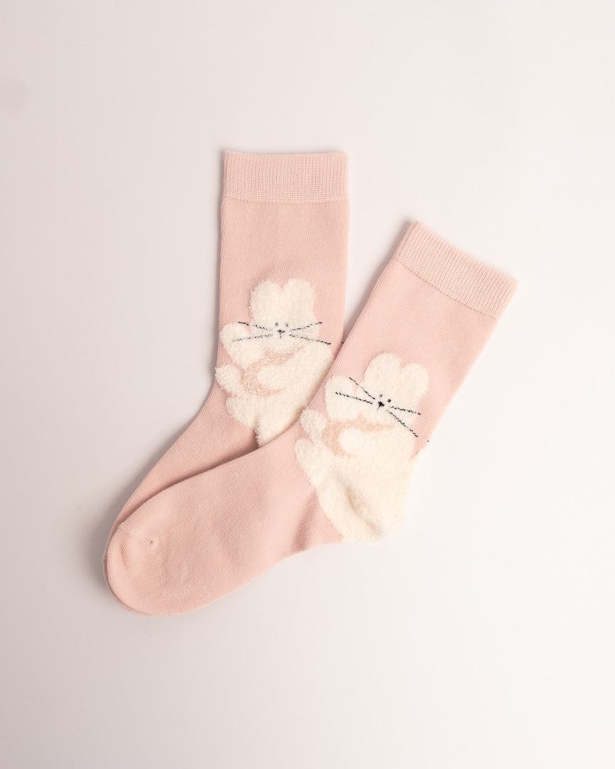 A pair of light pink crew socks featuring fluffy white bunny appliques on the front with embroidered black whiskers, displayed on a clean off-white background.