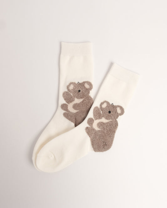 A pair of off-white crew socks featuring a textured, fuzzy brown koala design on the side, displayed on a clean, light gray background.