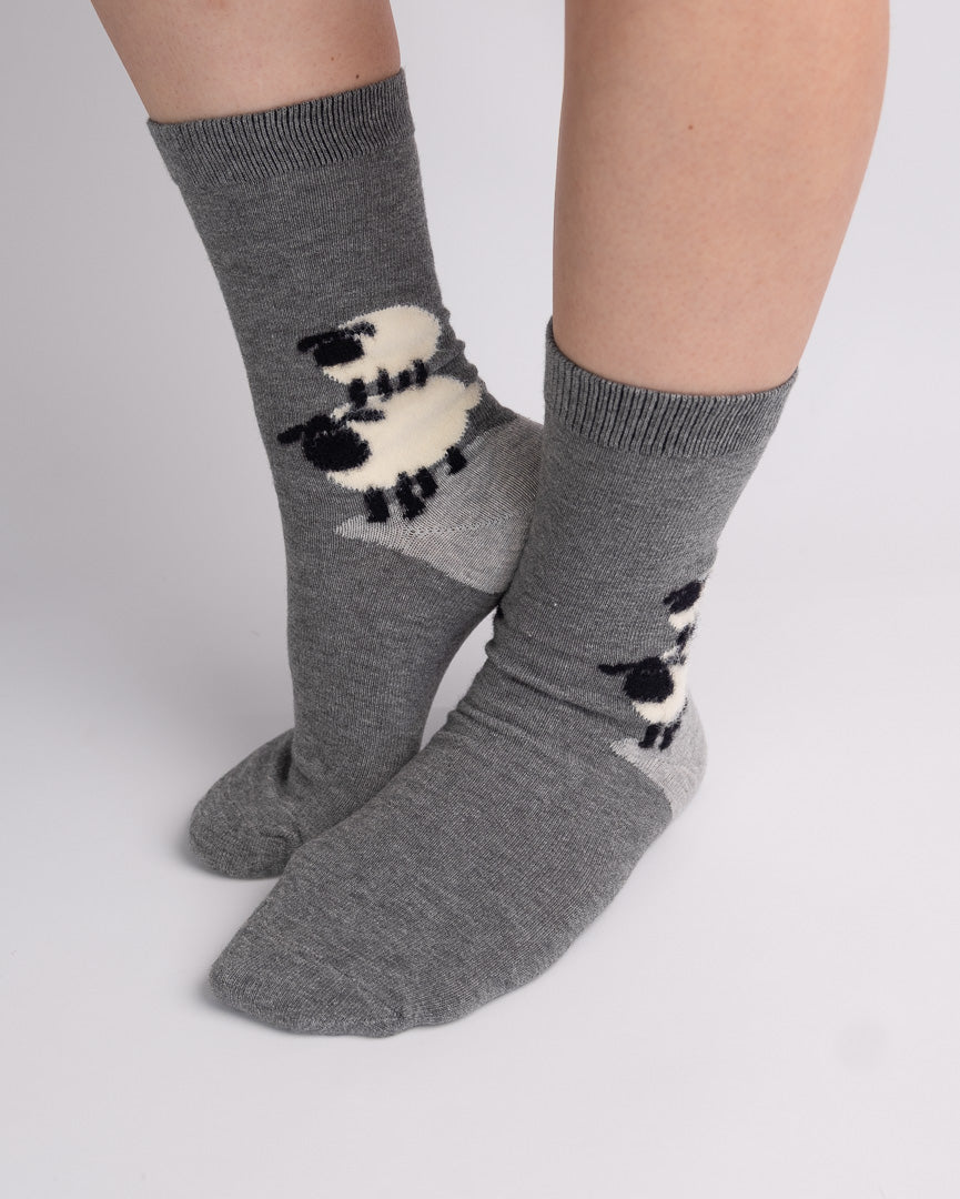 A pair of crew socks worn on feet, featuring a gray knit base with a black and white sheep pattern on the sides, shown against a light gray studio background.