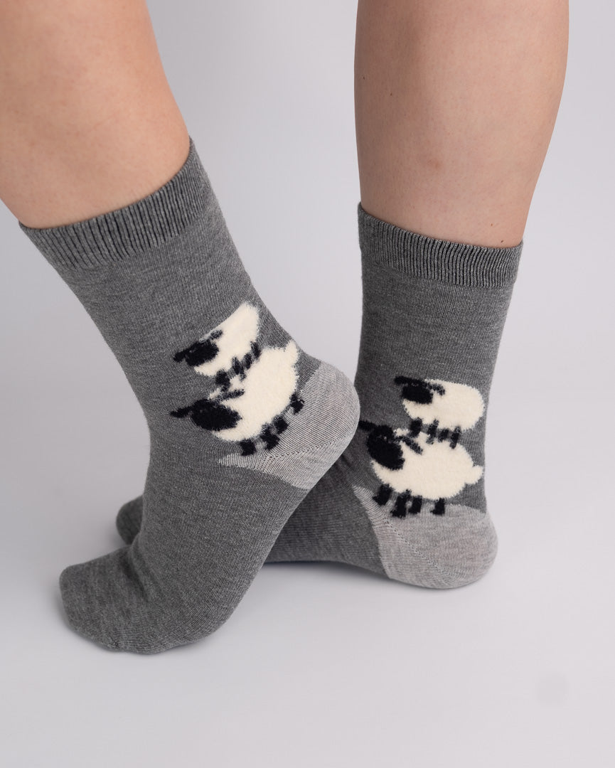 A pair of medium gray crew socks with a black and white sheep pattern, shown on a person's feet against a light gray studio background.