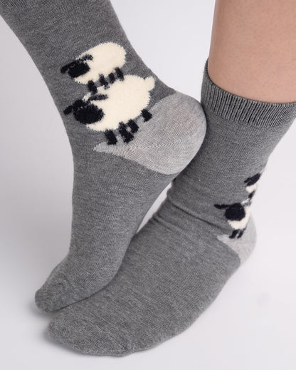 A pair of gray crew socks with a black and white sheep pattern, shown on a person's feet against a light gray background.