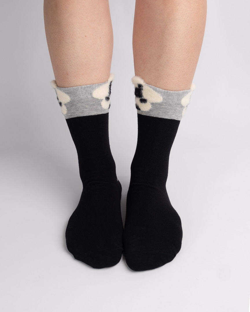 Black crew socks with a gray and cream knit floral cuff, shown on a person's feet against a light gray studio background.