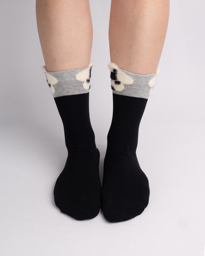 Black crew socks with a gray and cream knit floral cuff, shown on a person's feet against a light gray studio background.