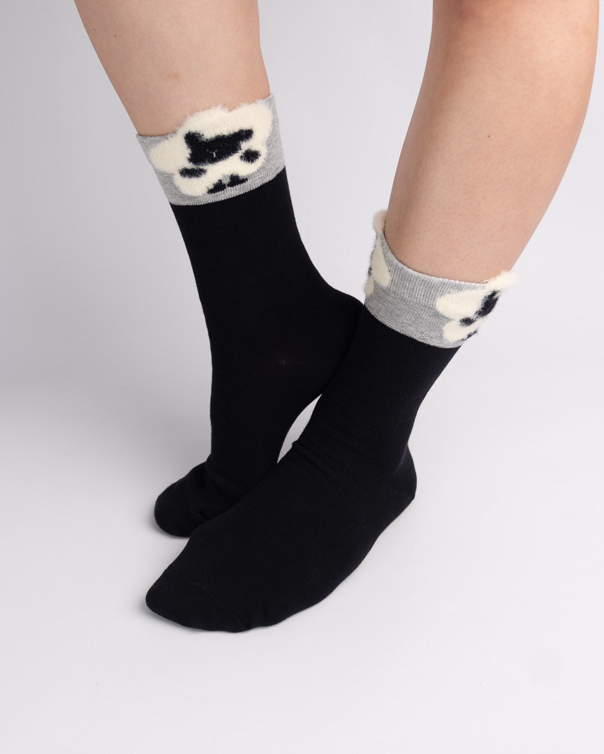 Black crew socks with gray fuzzy trim and a black-and-white paw print pattern, shown on a person's feet against a light gray studio background.