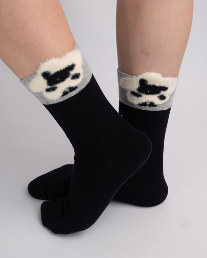 A pair of black crew socks with a fuzzy white and gray cuff featuring a black cat silhouette, shown on a person's feet against a light gray background.