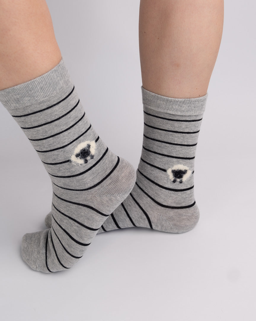 A pair of gray crew socks with black horizontal stripes and small black-and-white sheep motifs near the lower calf, shown worn on a person's feet against a light gray studio background.