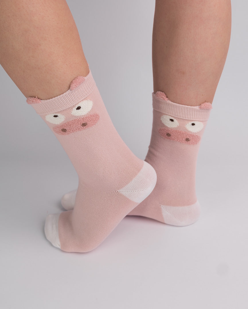A pair of light pink crew socks with a cute cartoon pig face design on the cuff, shown on a person's feet against a light gray studio background.