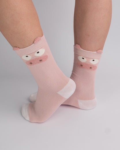 A pair of light pink crew socks with a cute cartoon pig face design on the cuff, shown on a person's feet against a light gray studio background.