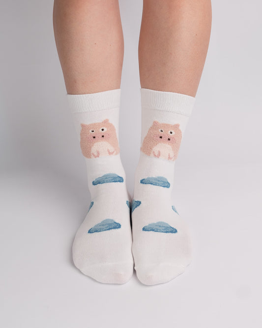 A pair of white crew socks worn on a person's feet, featuring a cute pink cartoon bear sitting on a blue cloud pattern.