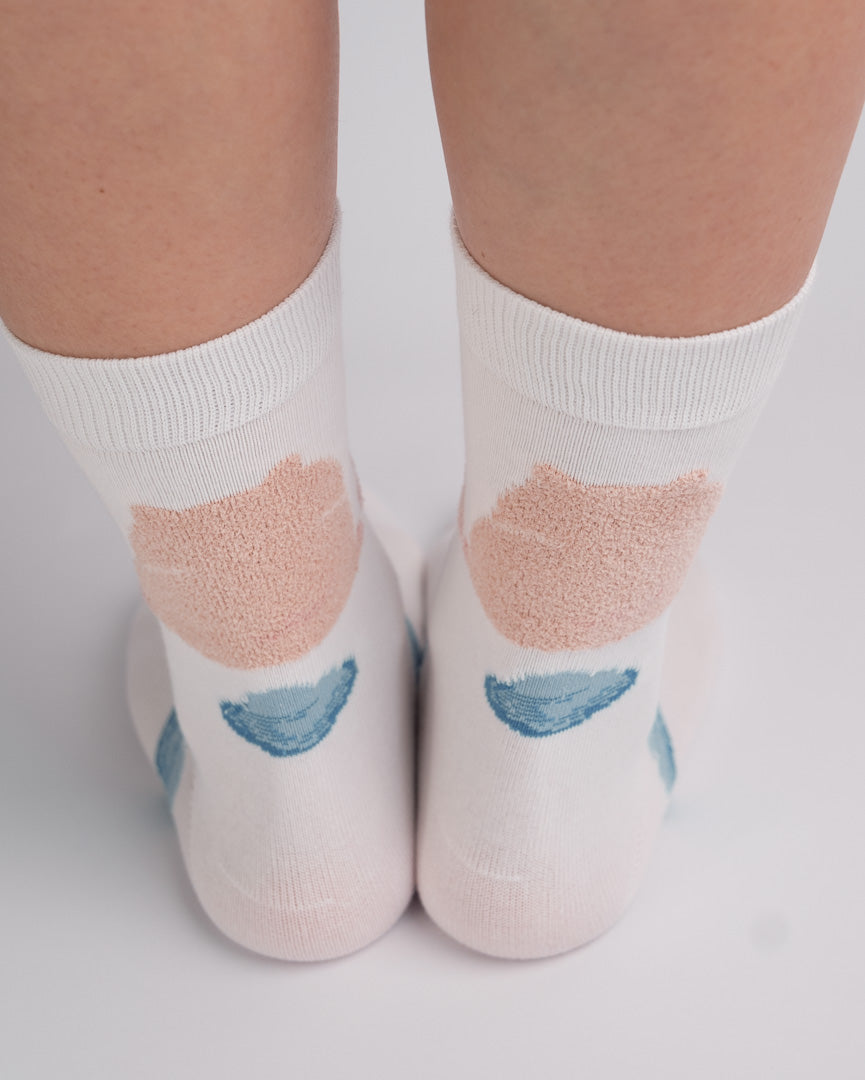 White ankle socks with a textured peach-pink heart and a blue crescent moon design, shown on a person's feet against a light gray background.