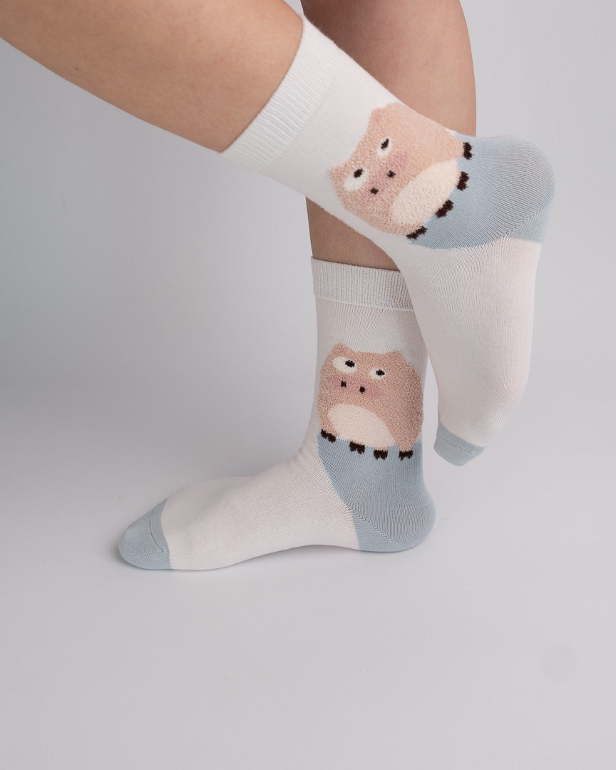 White crew socks with a light blue heel and toe and a pink cartoon animal face graphic, shown worn on a person's feet against a light gray studio background.