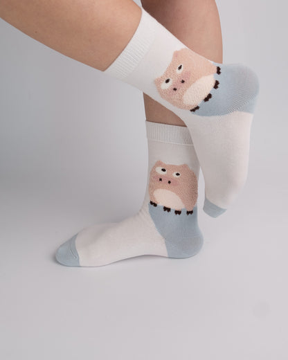 White crew socks with a light blue heel and toe and a pink cartoon animal face graphic, shown worn on a person's feet against a light gray studio background.