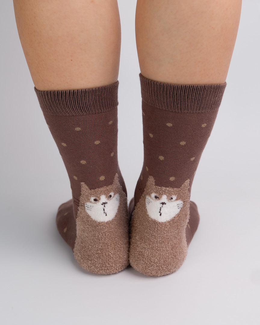 A pair of brown crew socks worn on feet, featuring a small cat face design on the front and small tan polka dots on a medium brown background.