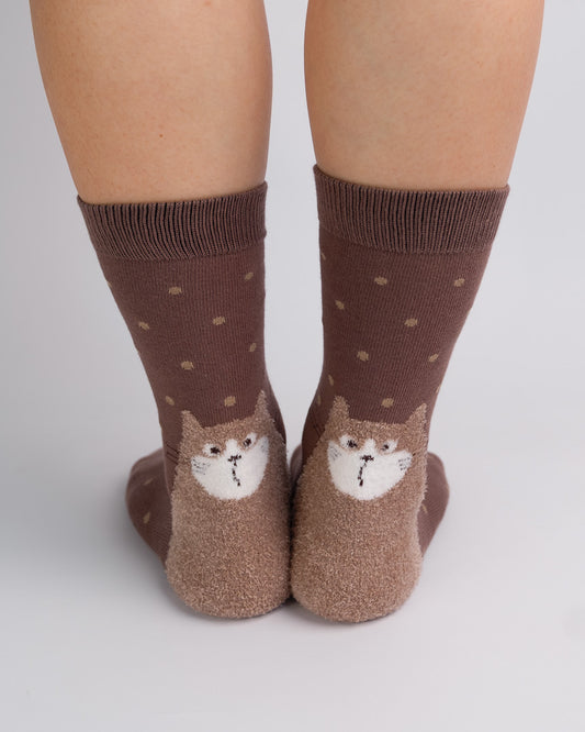 A pair of brown crew socks worn on feet, featuring a small cat face design on the front and small tan polka dots on a medium brown background.