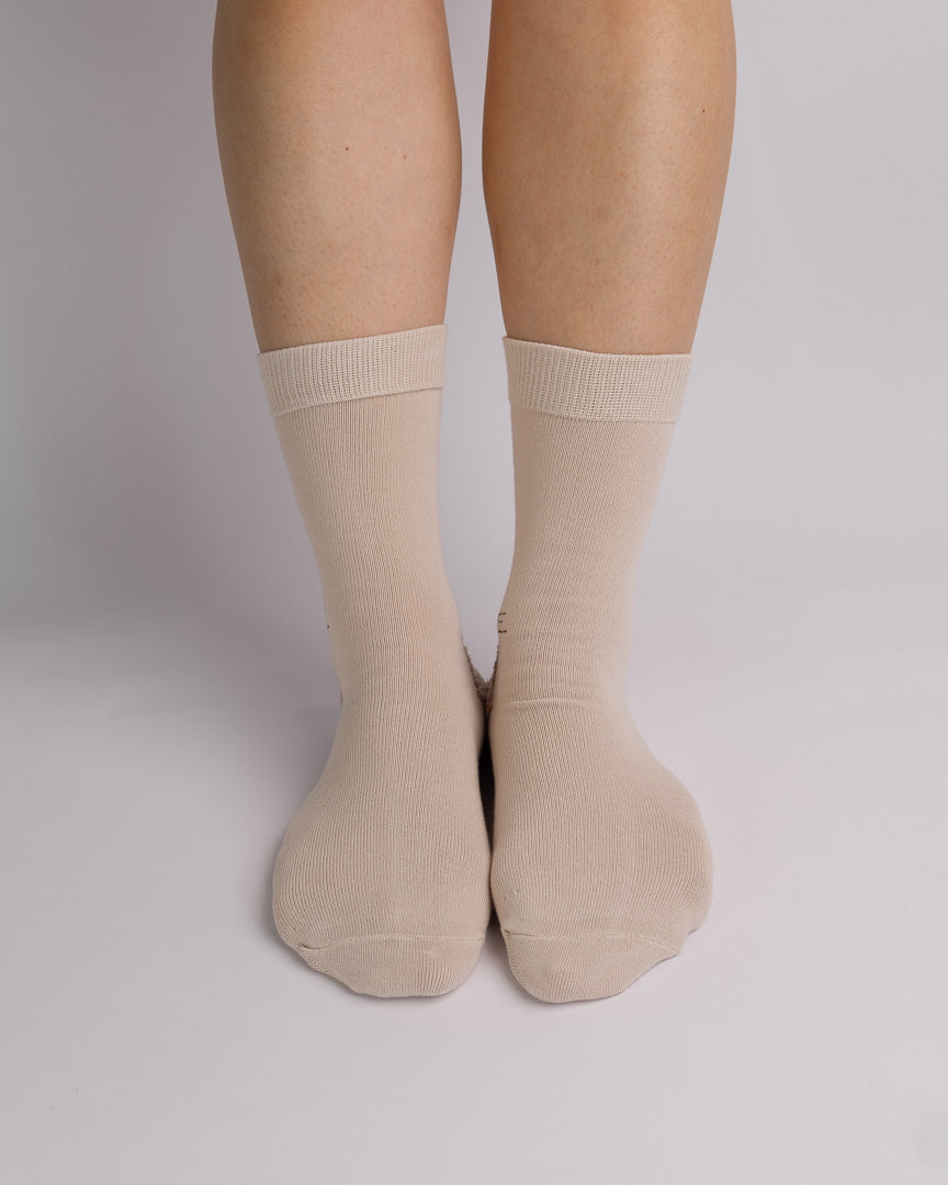 A pair of crew-length ribbed socks in a light beige color, shown on a person's feet against a plain light gray studio background.