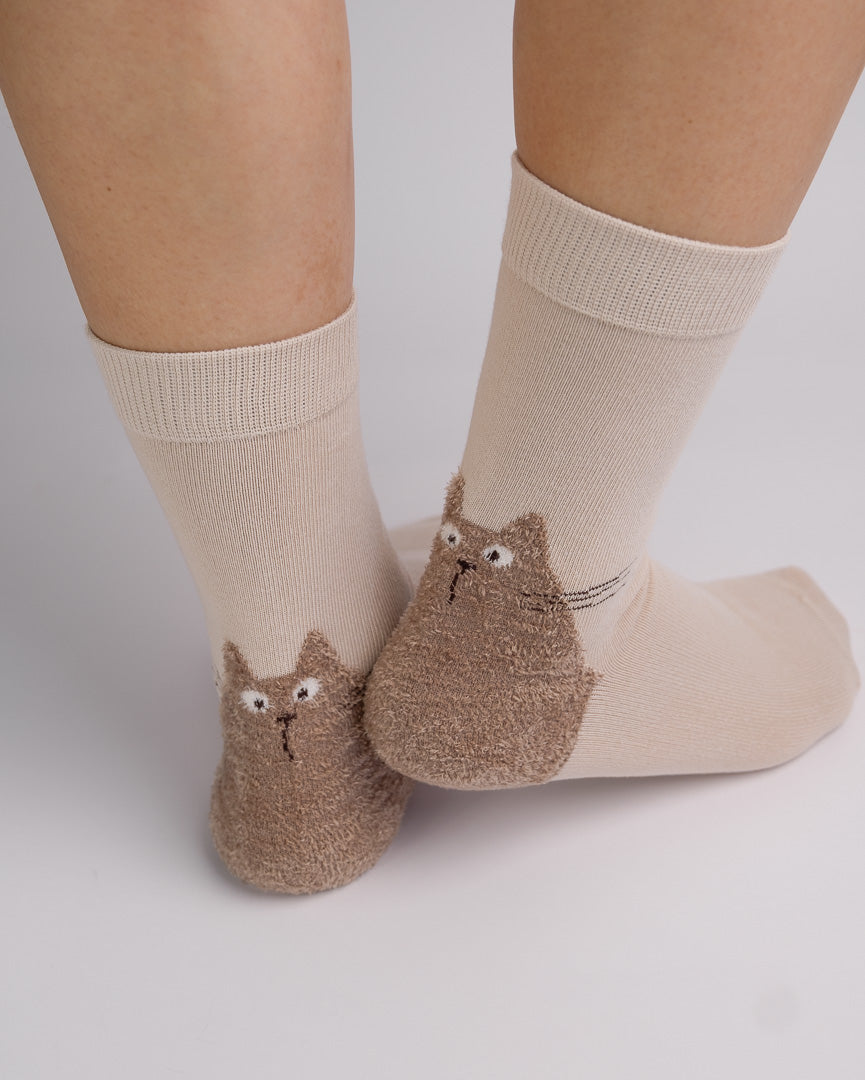 A pair of beige crew socks with brown fuzzy cat-shaped heels, shown on a person's feet against a light gray background.