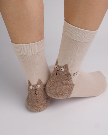 A pair of beige crew socks with brown fuzzy cat-shaped heels, shown on a person's feet against a light gray background.
