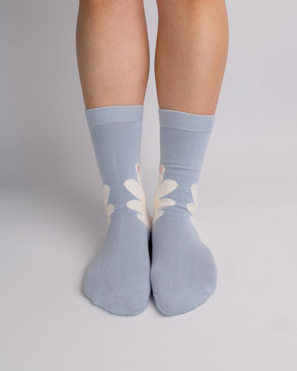 A pair of light blue crew socks with a white daisy pattern, worn on a person's feet against a neutral gray studio background.