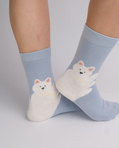 A pair of light blue crew socks with a white fluffy cat graphic on the front, worn on a person's feet against a light gray studio background.