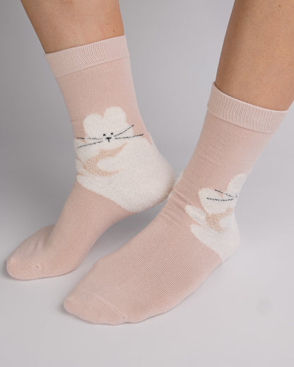 A pair of light pink crew socks with a white fuzzy bunny graphic on the side, shown on a person's feet against a neutral gray background.