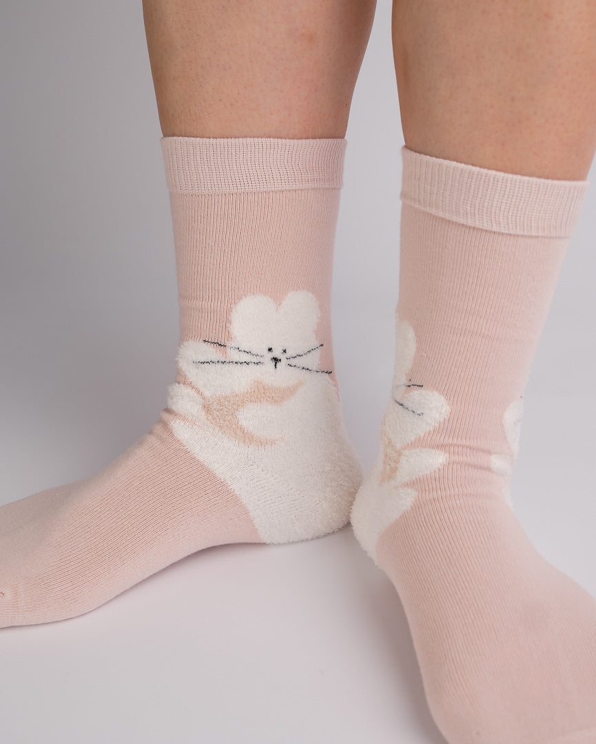 A pair of light pink crew socks with a white fluffy cat design on the side, shown worn on a person's feet against a neutral light gray background.