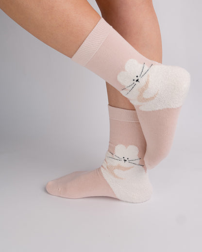 A pair of light pink and white fuzzy socks with a cat face design, shown on a person's feet against a light gray studio background.