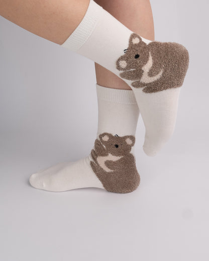 A pair of off-white crew socks with brown and cream knit animal footpads shaped like bears, shown on a person's feet against a light gray studio background.