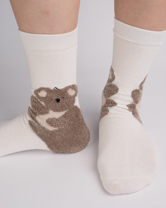A pair of off-white crew socks with a brown fuzzy bear graphic on the sole and heel, shown on a person's feet against a light gray background.