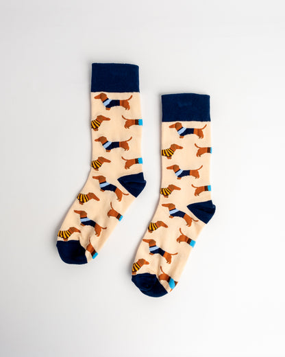 A pair of crew socks with a cream background featuring a pattern of dachshunds wearing sweaters and hats, with navy blue cuffs, heels, and toes, displayed on a light gray background.