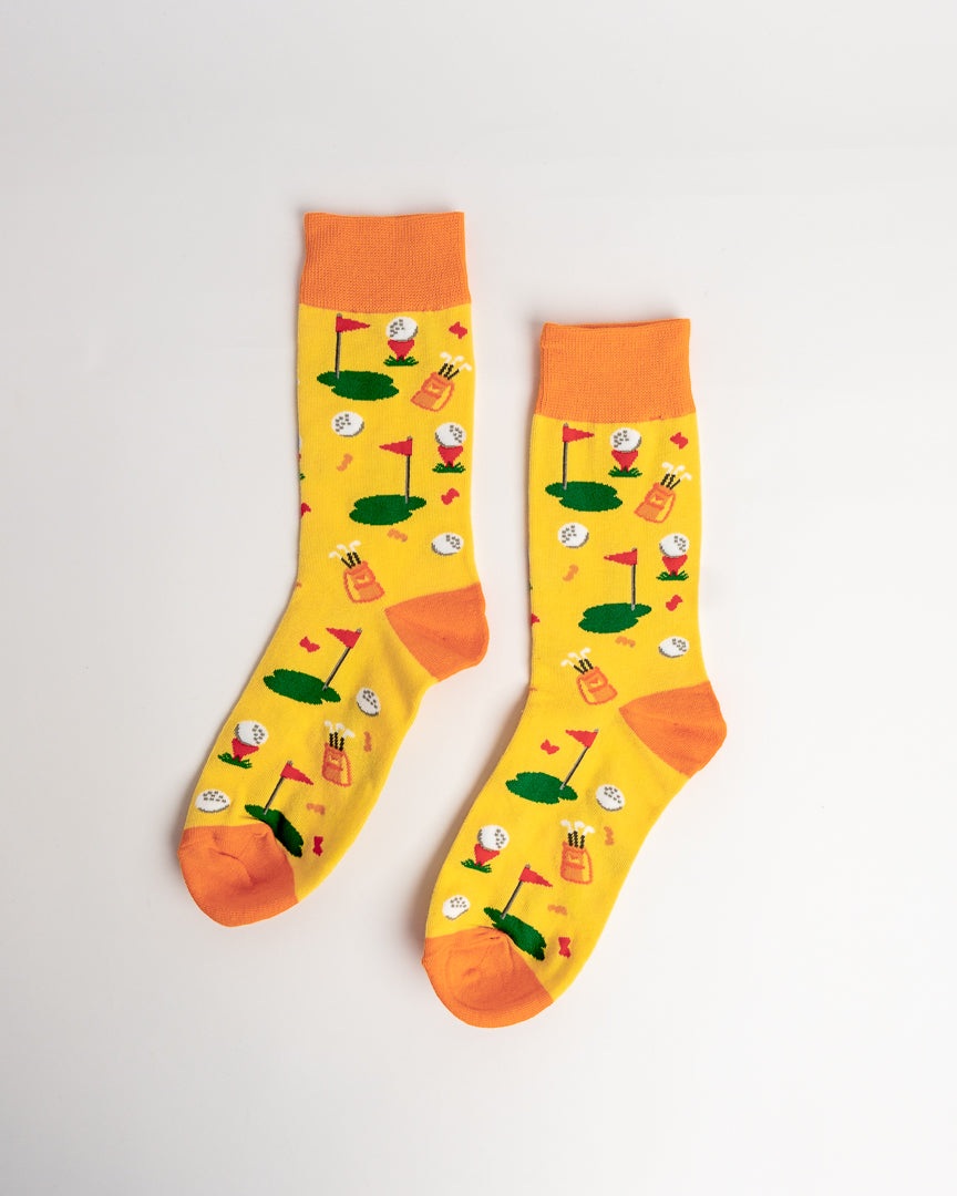 A pair of bright yellow crew socks with orange cuffs, heels, and toes, featuring a playful golf-themed print of mini golf balls, flags, and greens.