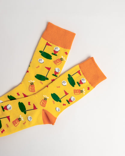 A pair of bright yellow crew socks with orange cuffs and toes, featuring a playful golf course pattern with green tees, flags, and golf balls, displayed on a clean white background.