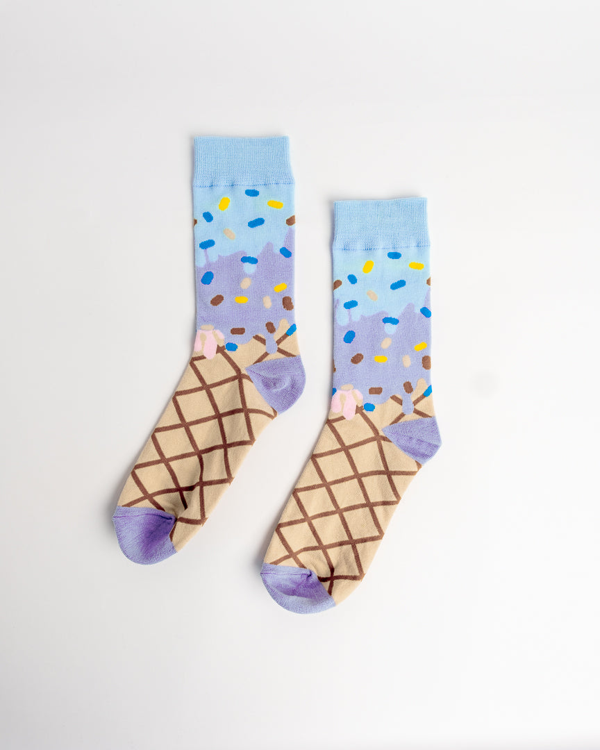 A pair of novelty socks featuring a printed ice cream cone design. The lower half of the socks depicts a waffle cone with brown lattice lines, while the upper half shows a light blue background with purple ice cream, yellow, blue, and brown sprinkles, and a small pink swirl.