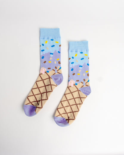 A pair of novelty socks featuring a printed ice cream cone design. The lower half of the socks depicts a waffle cone with brown lattice lines, while the upper half shows a light blue background with purple ice cream, yellow, blue, and brown sprinkles, and a small pink swirl.